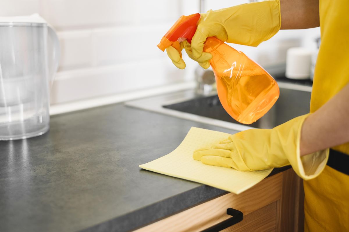 Person in yellow gloves spraying and wiping a dark kitchen countertop